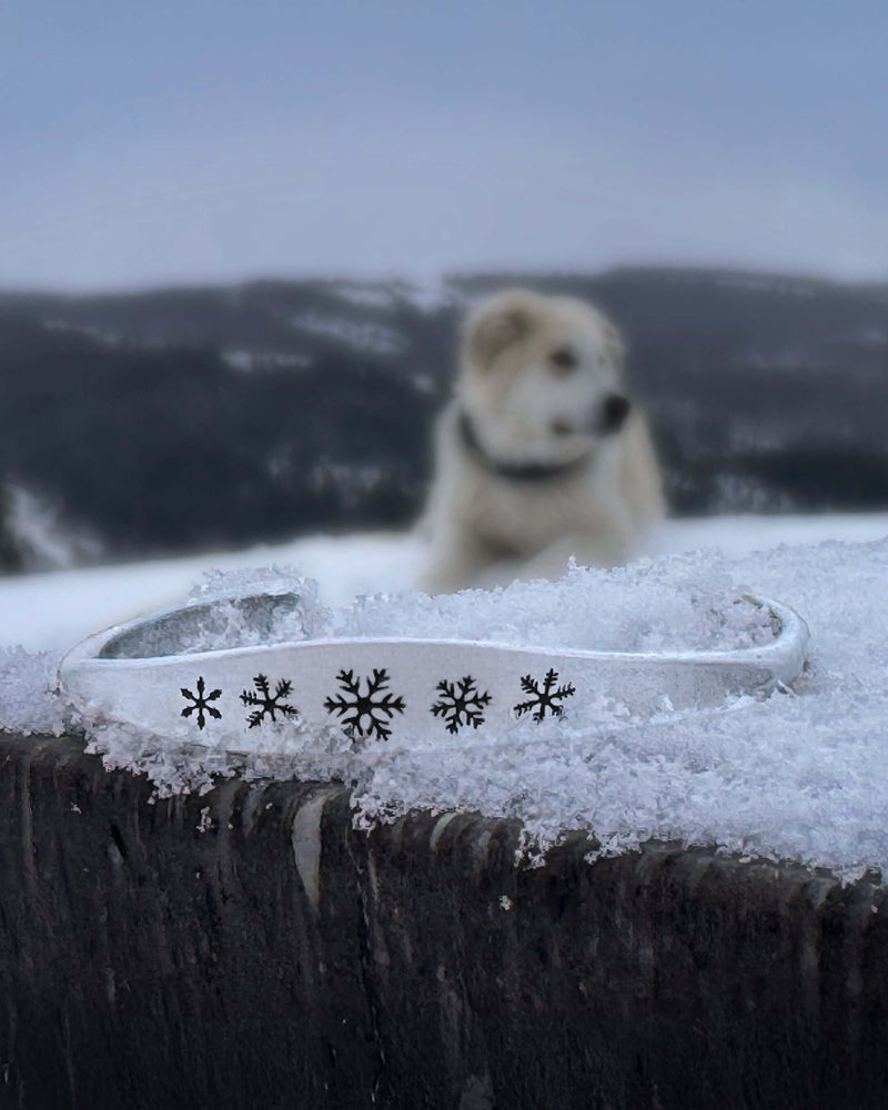 Powder Signet Cuff Bracelet resting in snow with snowflakes design and a dog in the background.