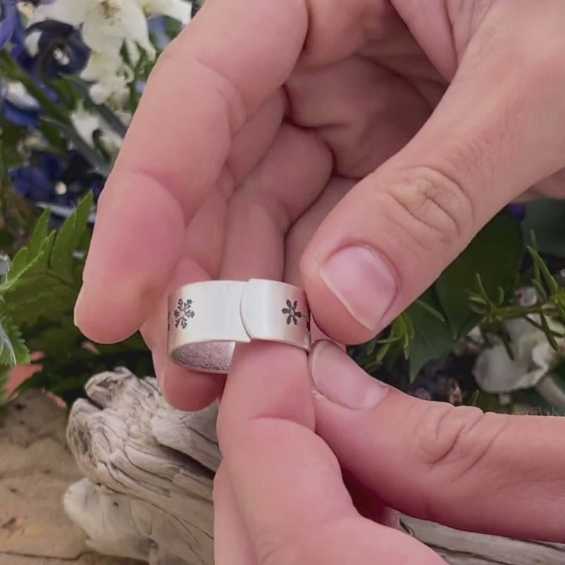 Person holding the Snowfall Adjustable Ring with etched snowflakes, surrounded by greenery and floral background.
