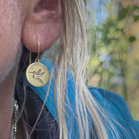 Close-up of antique brass hummingbird earrings dangling from a woman's ear, showcasing detailed engraving.