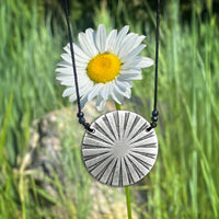Ray Necklace hanging in front of a daisy, showcasing its sun ray design and matte silver finish.