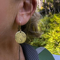 Close-up of Rising Wolf Earrings featuring a topographic design, worn by a person outdoors in nature.