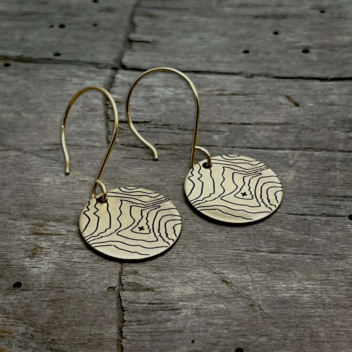 Rising Wolf Earrings featuring topographic design in raw brass with gold-filled earwires on a rustic wood background.