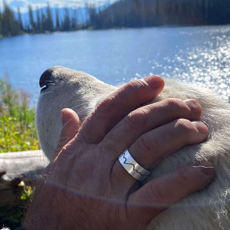 A hand wearing a Simple Mountain Ring rests on a dog's head by a lakeside view.