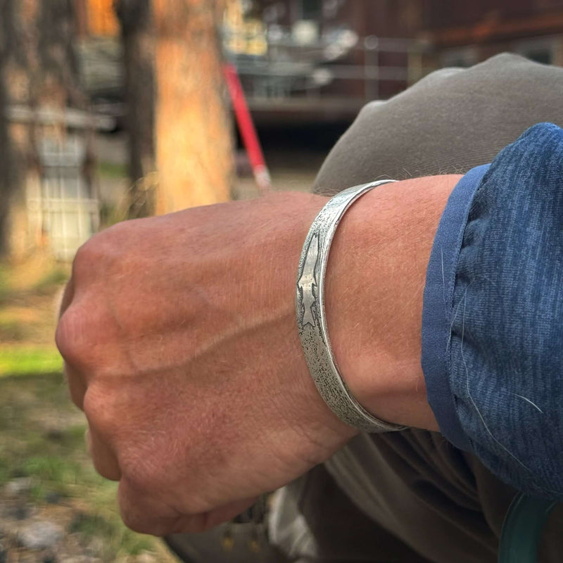 Close-up of a hand wearing a Single Trout Cuff Bracelet outdoors, showcasing the adjustable design and trout engraving.