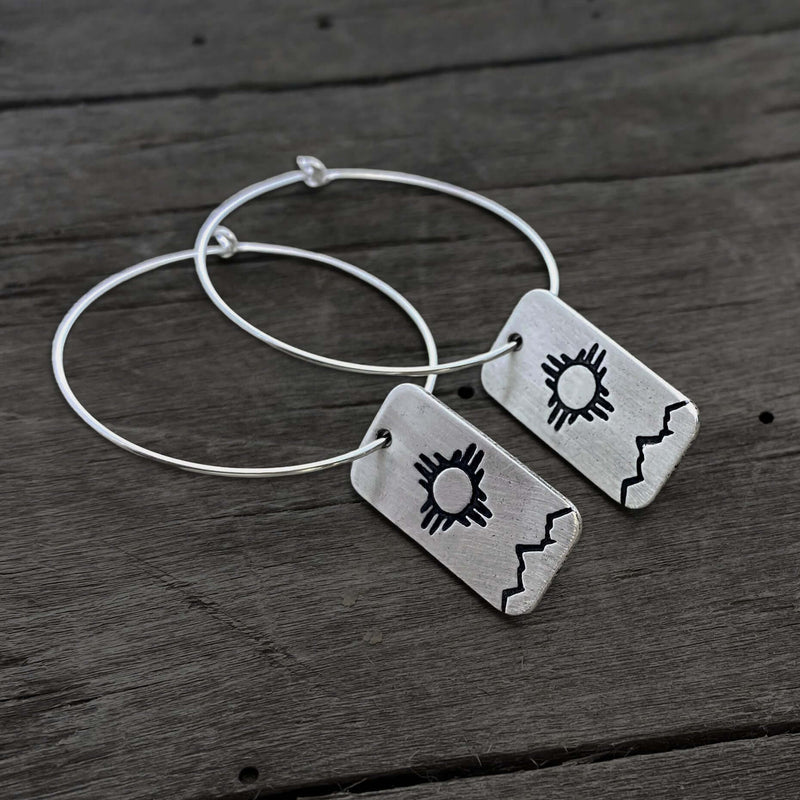 Sol Hoop Earrings featuring Zia sun design in raw brass and sterling silver finish on wooden background.