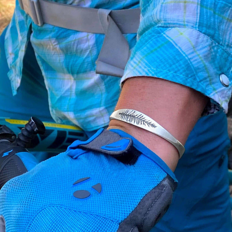 Person wearing a blue glove showcasing the Spruce Signet Cuff Bracelet with a spruce outline etching.