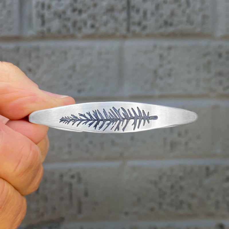 Hand holding the Spruce Signet Cuff Bracelet with intricate spruce outline etching.