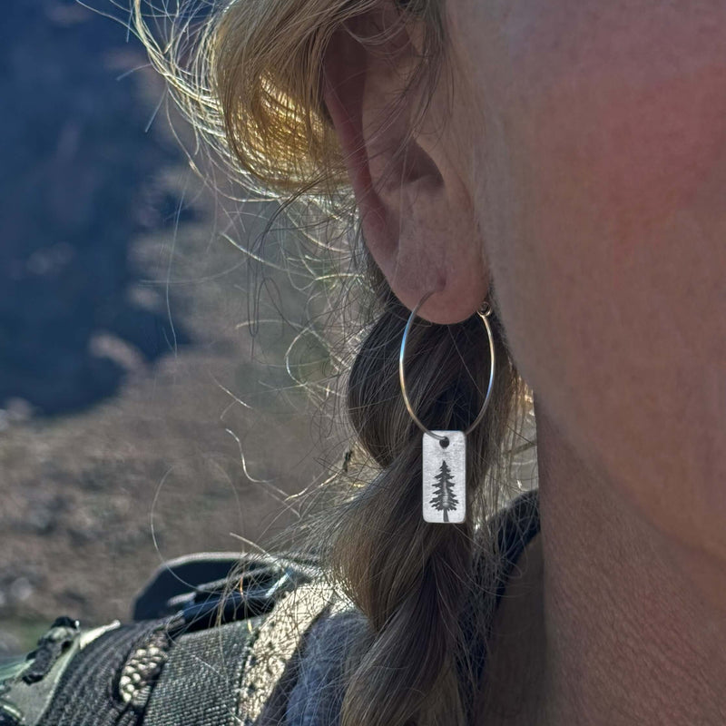 Close-up of Spruce Tree Hoop Earrings on a woman's ear with a scenic background.