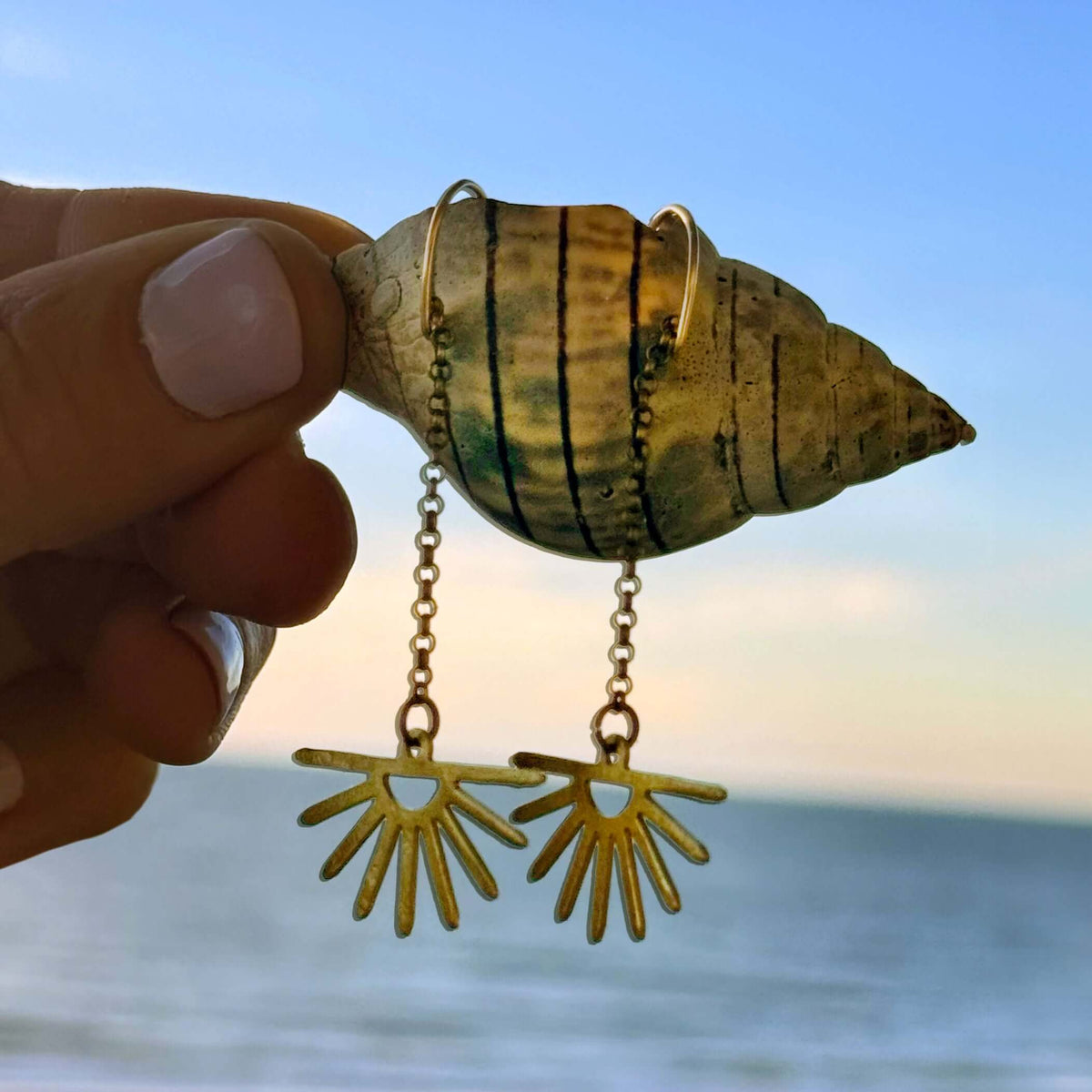 Sunbeam Earrings displayed on a shell against a beach sunset, showcasing their radiant design and delicate chains.