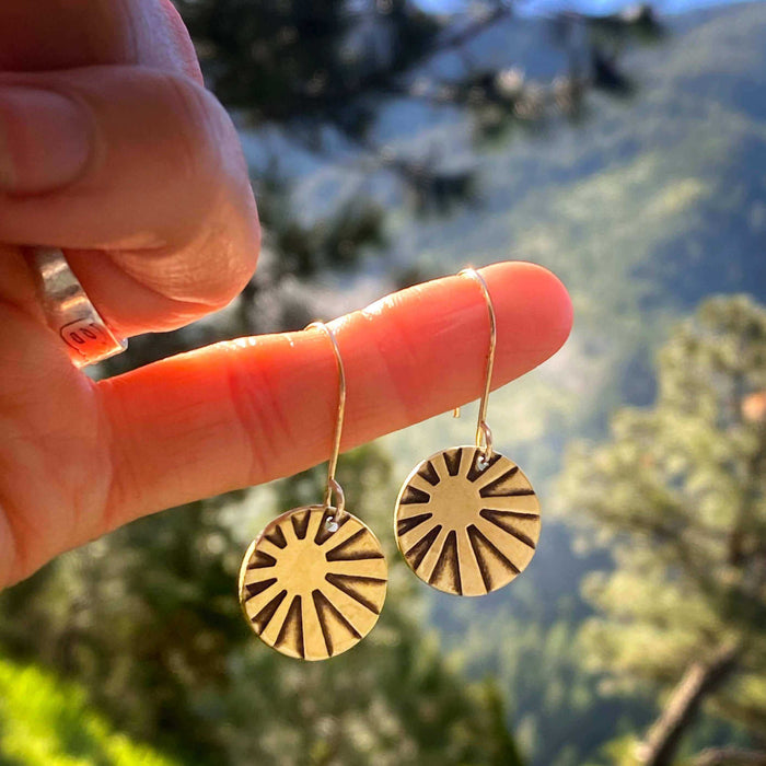 Mini Sun Ray  Hoop Earrings held between fingers in a scenic outdoor setting, featuring a sun ray design in raw brass.