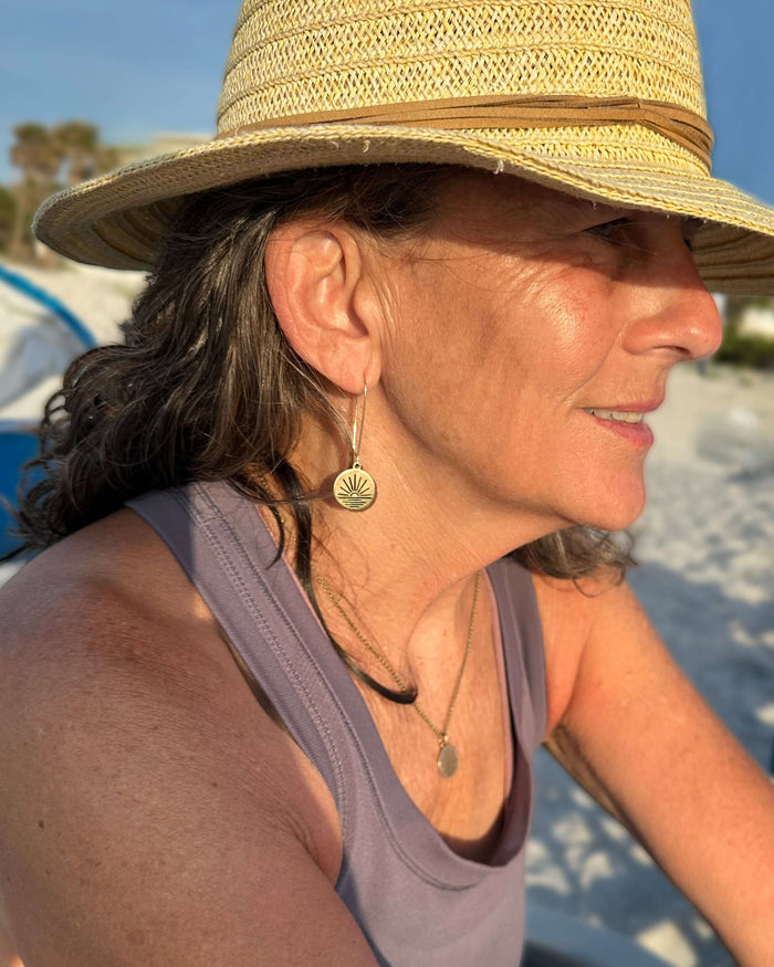 Woman wearing Sunrise Earrings with a simple sunrise design, enjoying a beach day in a straw hat.