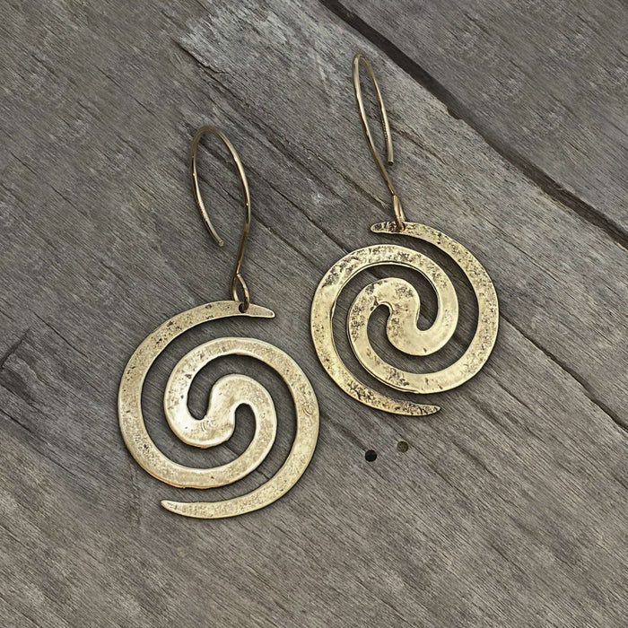 Vortex Earrings in antique brass on wooden background, featuring lightly hammered spiral design.
