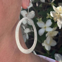 Wide Open Earrings in matte silver showcasing elegant texture and design, worn against a floral background.