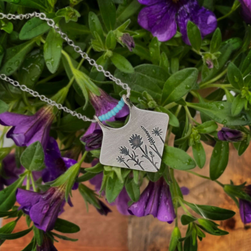 Wildflower Box Necklace featuring floral design and turquoise accent on a chain, surrounded by purple flowers.