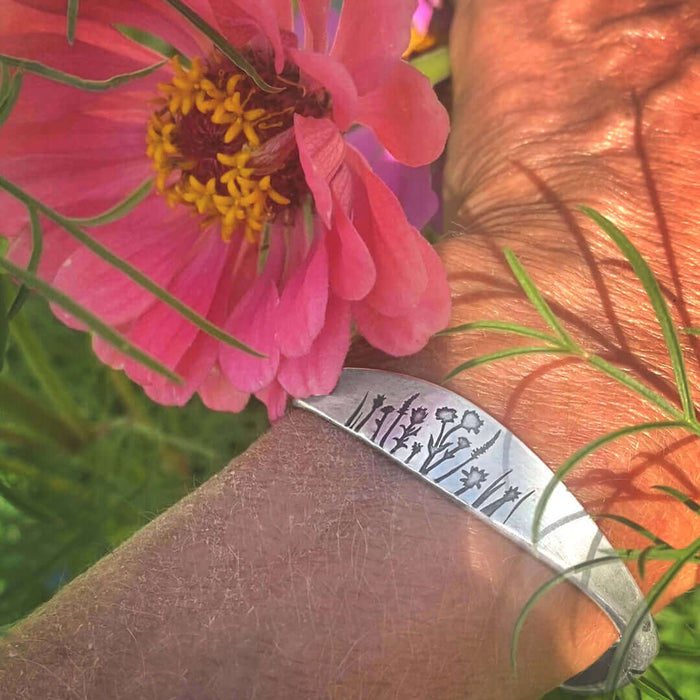 Wildflowers Signet Cuff adorned with floral engravings, worn with vibrant flowers in the background.