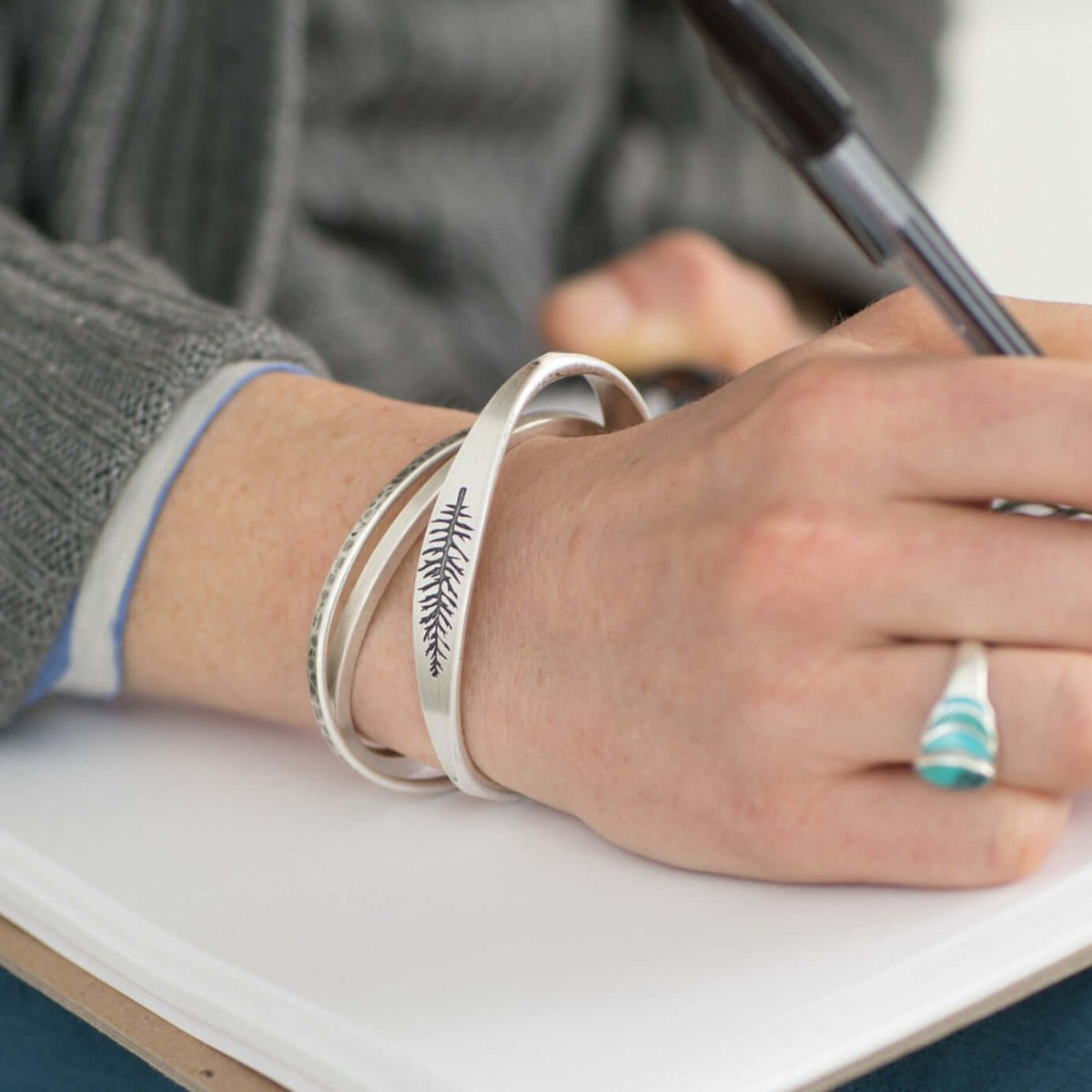 Person wearing three bracelets, including the All Square Cuff Bracelet, while writing in a notebook.