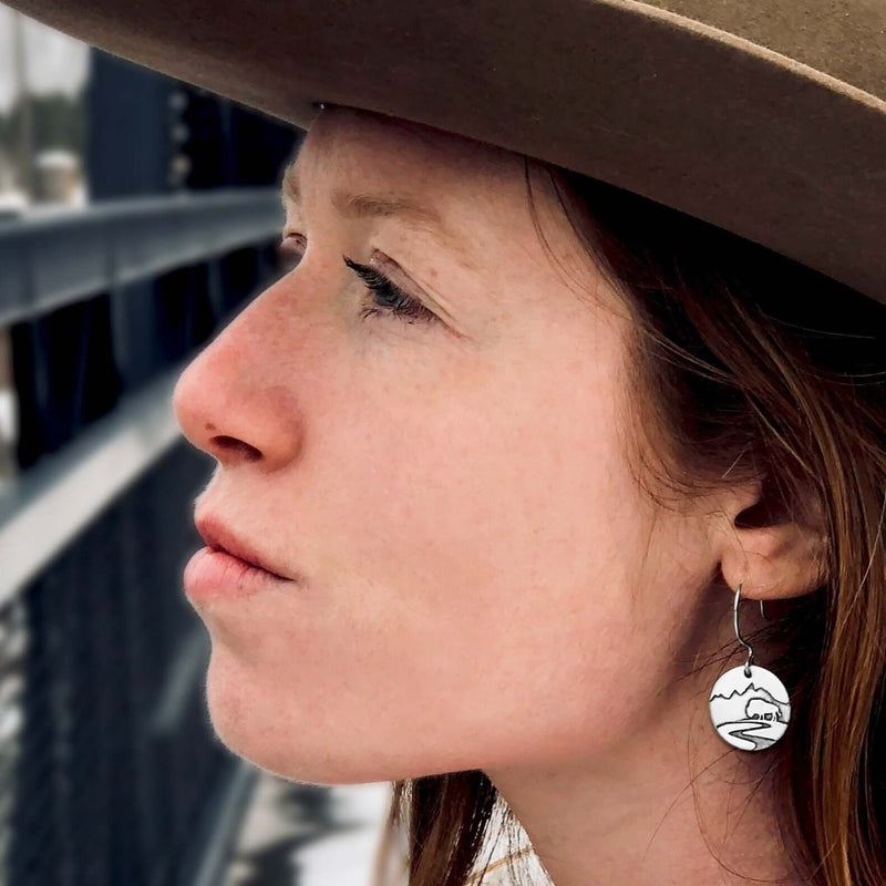 Buffalo in the Tetons Earring worn by a woman, showcasing the reflective buffalo and mountain design against a scenic backdrop.