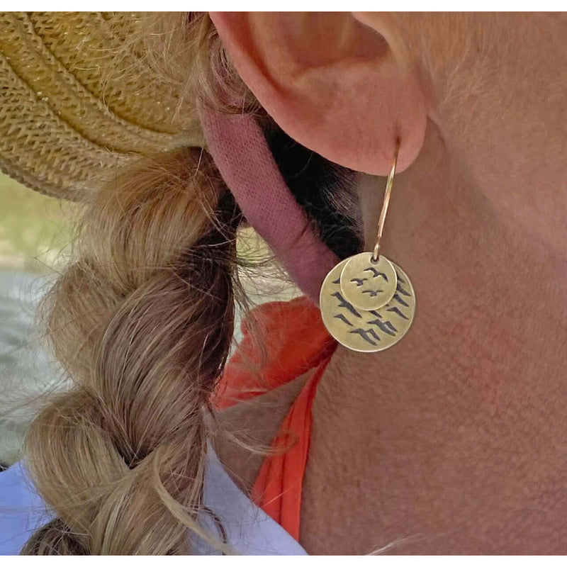 Woman wearing Flyover Earrings with beach-themed design featuring birds on a gold disk.