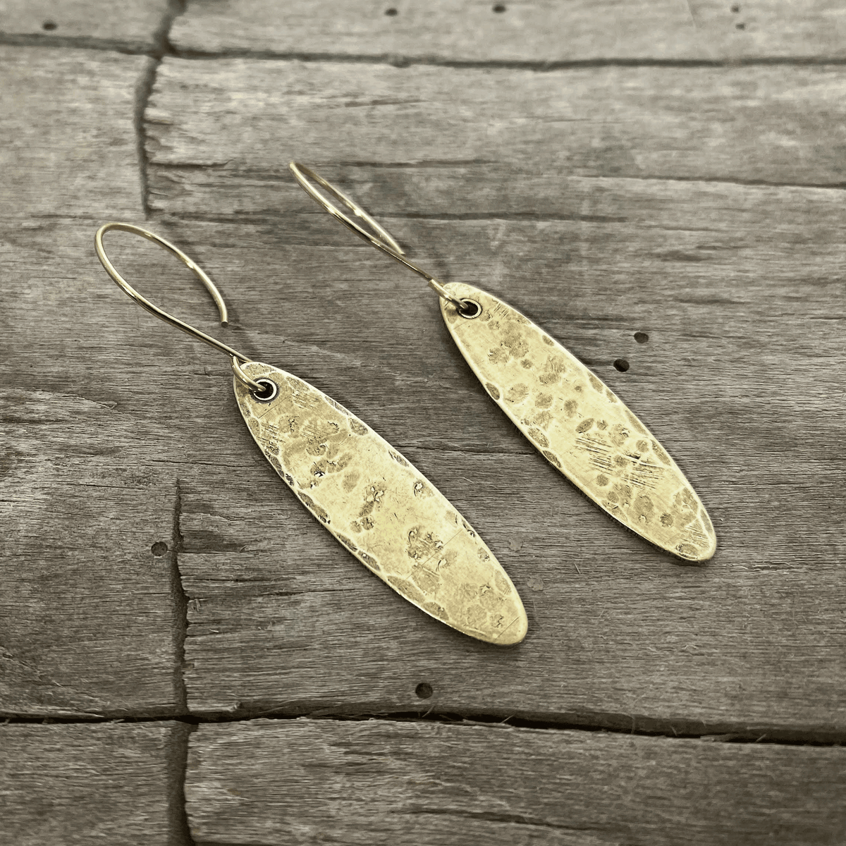 Fog Drop Earrings in antique brass finish with textured design hanging from earwires on a wooden background.
