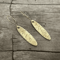 Fog Drop Earrings in antique brass finish with textured design hanging from earwires on a wooden background.
