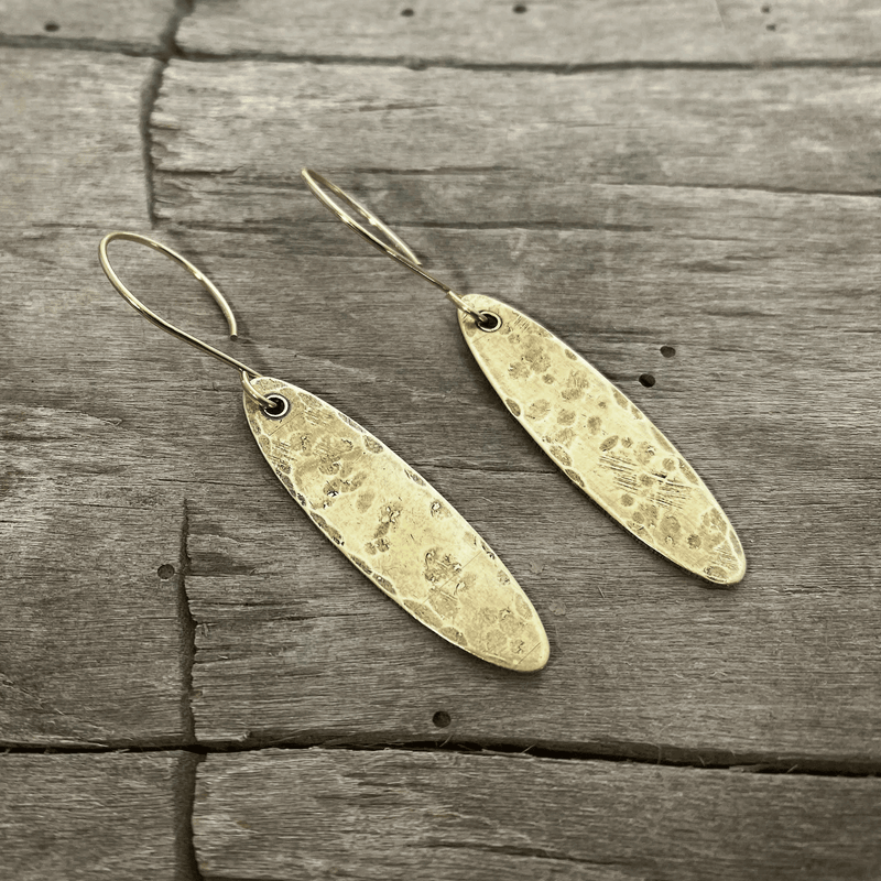 Fog Drop Earrings in antique brass finish with textured design hanging from earwires on a wooden background.