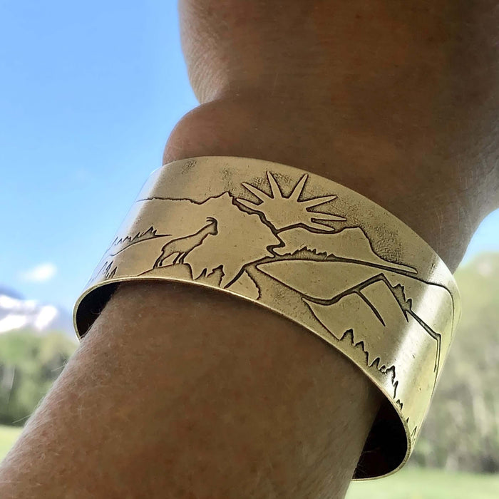 Going to the Sun Road Cuff Bracelet featuring mountain etched design in antique brass, worn on wrist with sunlight backdrop.