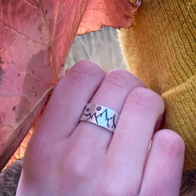 High Alpine Adjustable Ring worn on hand with mountain and star design, set against a colorful autumn background.
