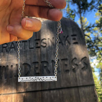 High Alpine Necklace showcased against a wilderness sign, featuring a scenic mountain design in silver.