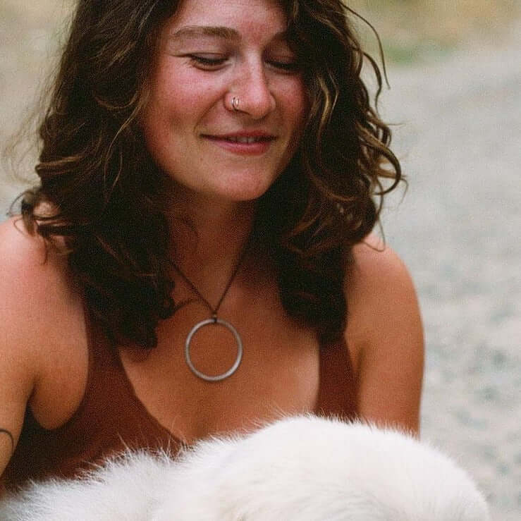 A woman wearing a Hula Necklace with a large circle hoop, smiling while petting a fluffy white puppy.
