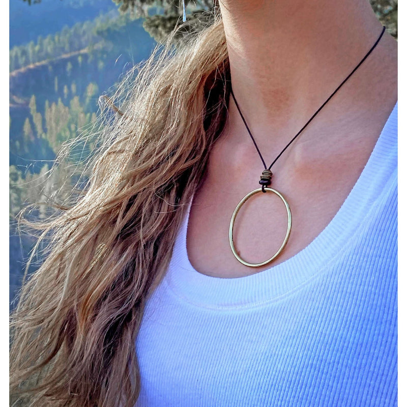 Hula Necklace featuring a raw brass circle hoop, adjustable slider cord, worn outdoors with natural landscape background.