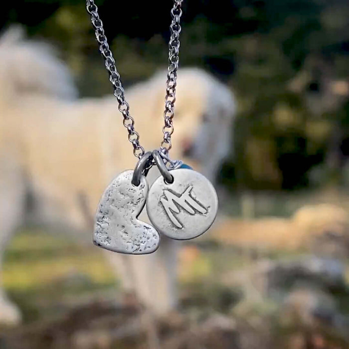 Close-up of the Love Montana Necklace featuring a heart and engraved MT disc against a blurred natural background.