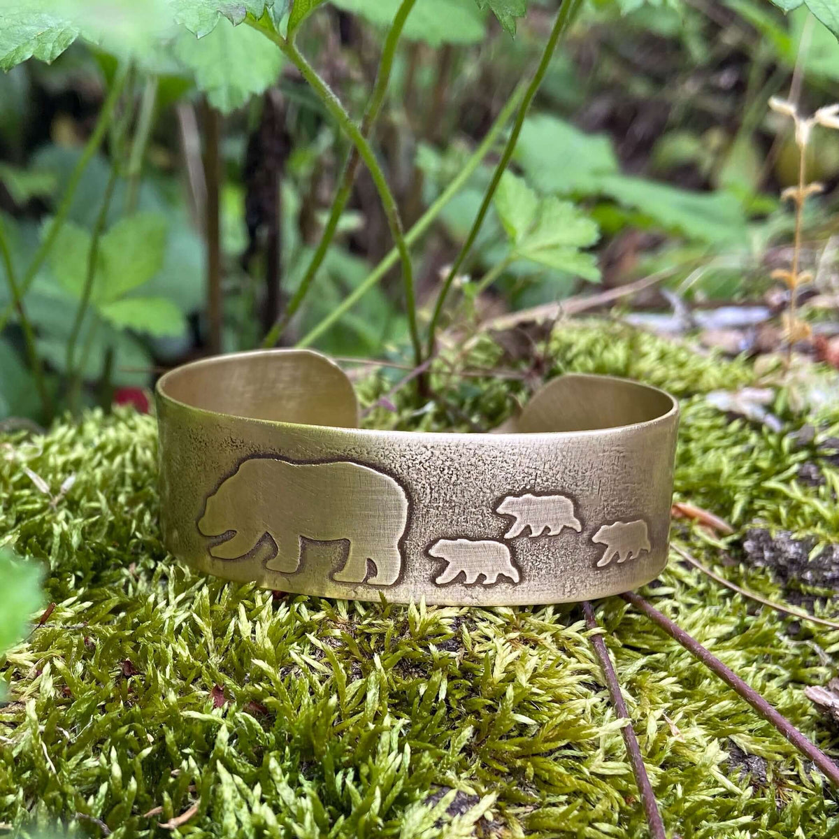 Mama and Cubs Cuff Bracelet featuring etched bear and cub designs on raw brass, surrounded by natural greenery.