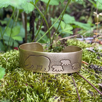 Mama and Cubs Cuff Bracelet featuring etched bear and cub designs on raw brass, surrounded by natural greenery.