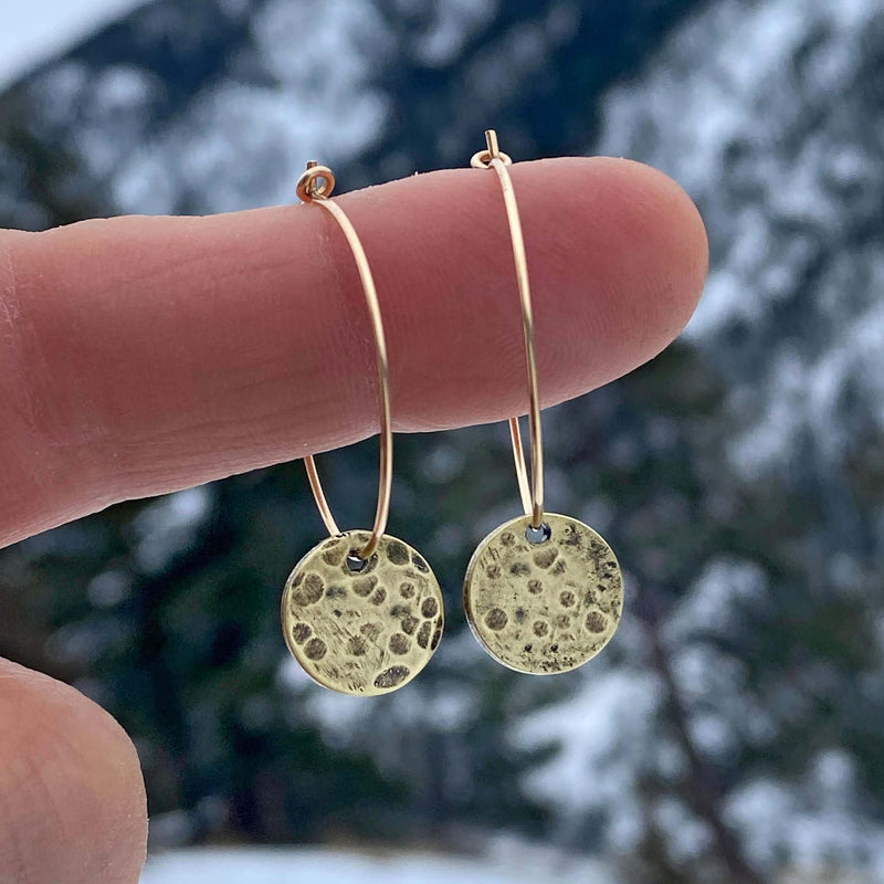 Mini Moon Hoop Earrings displayed on a finger against a scenic outdoor background.