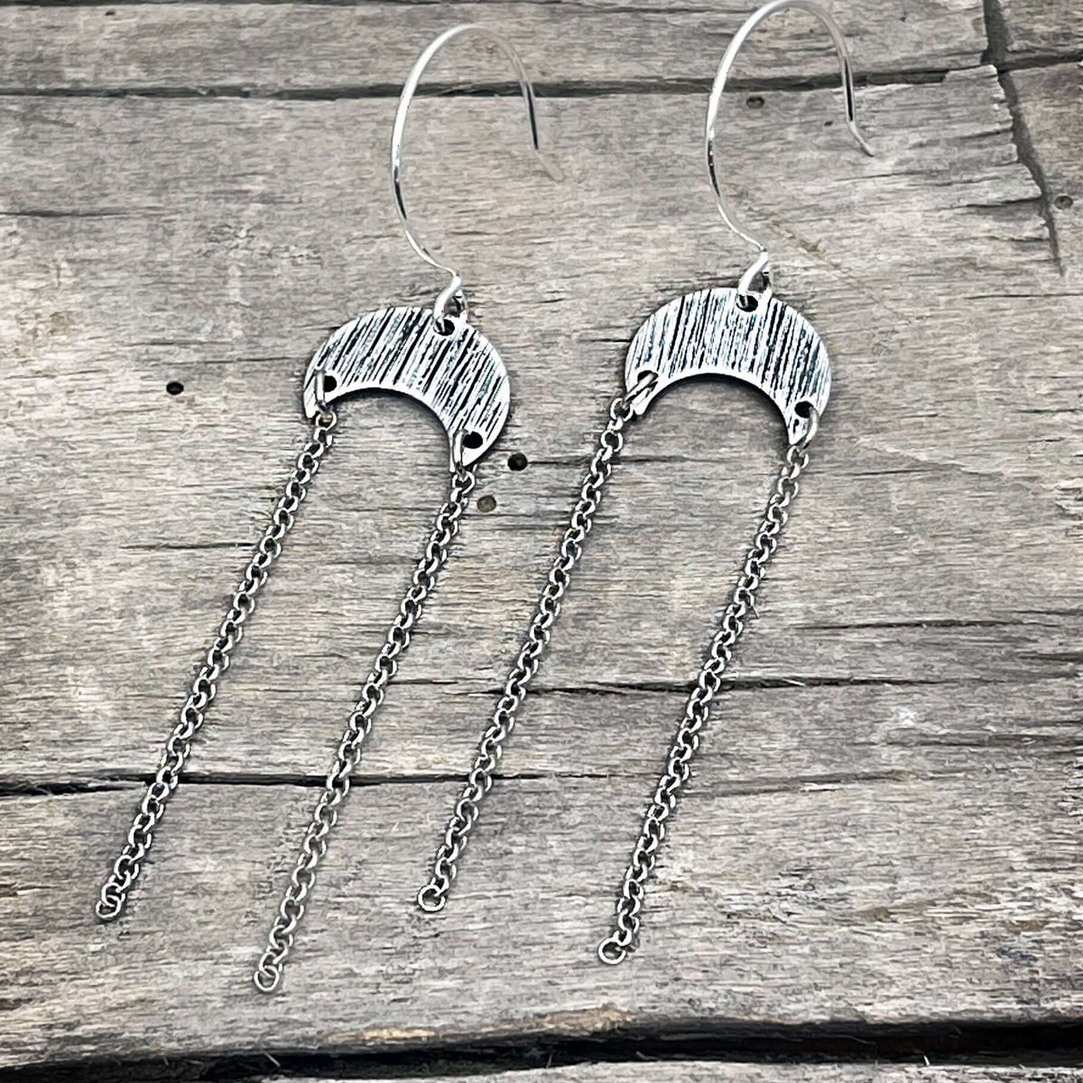 Moonfall Earrings in matte silver with dangly chains on a rustic wooden background.