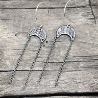 Moonfall Earrings in matte silver with dangly chains on a rustic wooden background.
