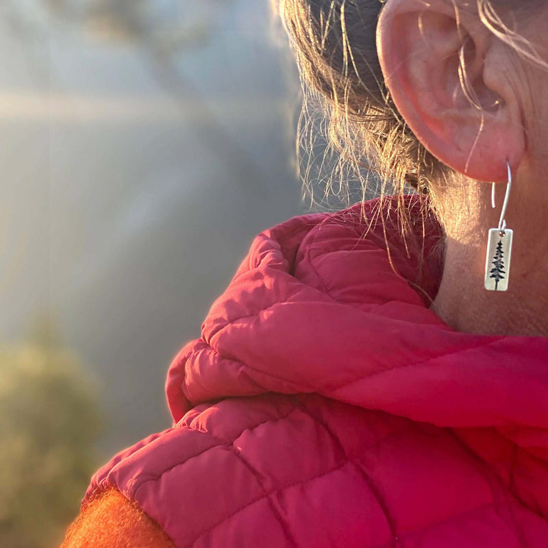 Close-up of a person wearing P Pine Tree Earrings, showcasing the design against a serene outdoor background.