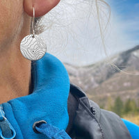 Close-up of Rising Wolf Earrings featuring topographic design worn outdoors with a scenic mountain backdrop.