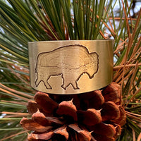 Roam Free Cuff in antique brass with buffalo engraving, set against pine and pinecone background.