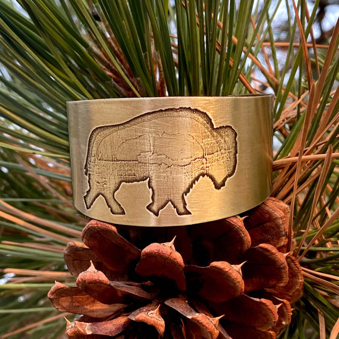 Roam Free Cuff in antique brass with buffalo engraving, set against pine and pinecone background.