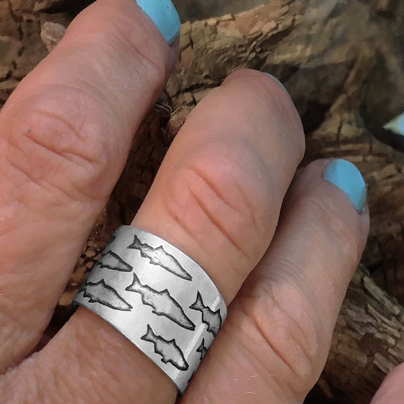 Salmon Run Adjustable Ring on a hand showcasing salmon etching design against natural wood background.