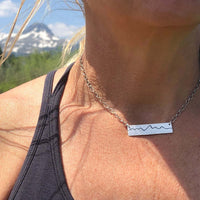 Simple Teton Mountain Necklace worn outdoors with mountain backdrop, showcasing the etched Teton range pendant.