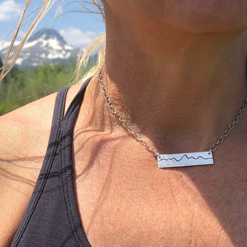 Simple Teton Mountain Necklace worn outdoors with mountain backdrop, showcasing the etched Teton range pendant.
