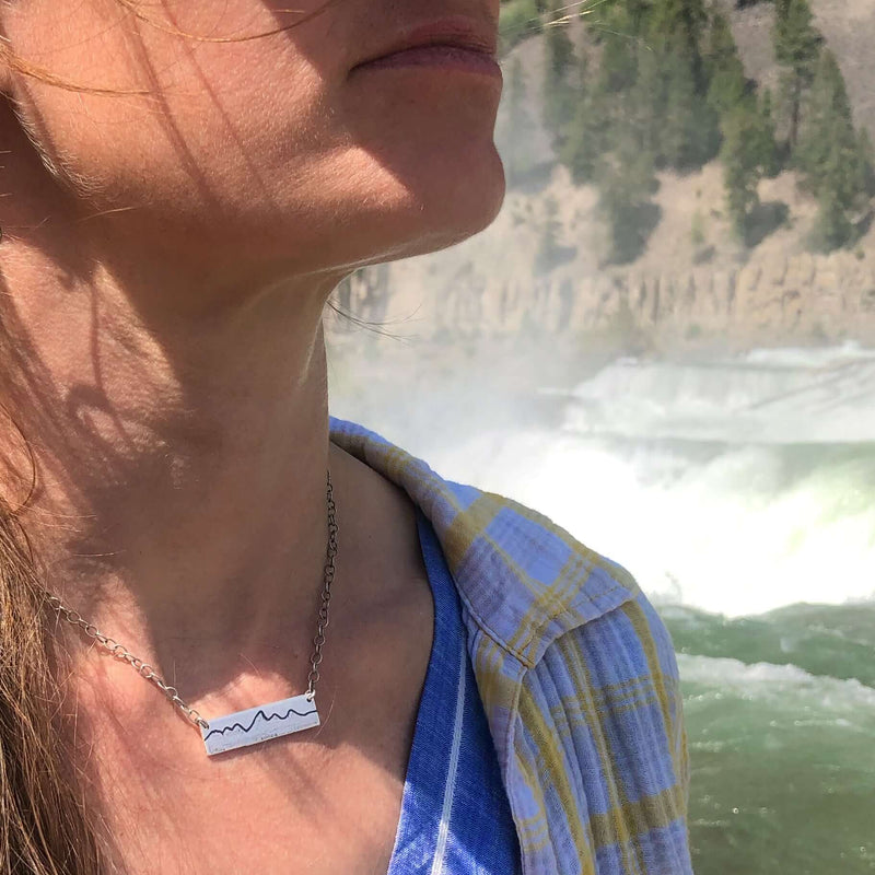 Simple Teton Mountain Necklace worn by a woman near a waterfall, showcasing the etched Teton mountain range design.