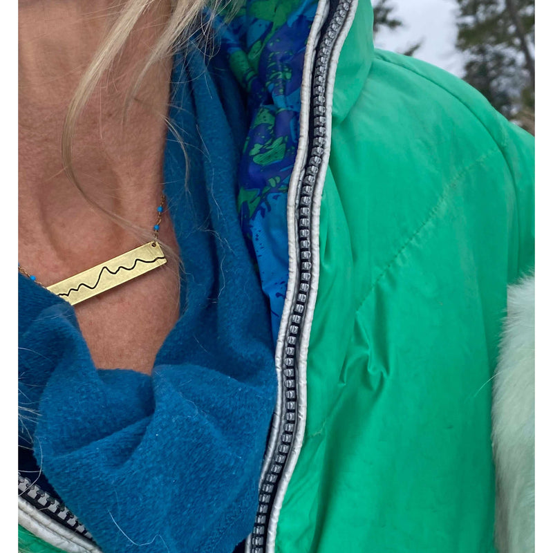 Woman wearing a Simple Teton Mountain Necklace with a green jacket and blue scarf, showcasing mountain range design.
