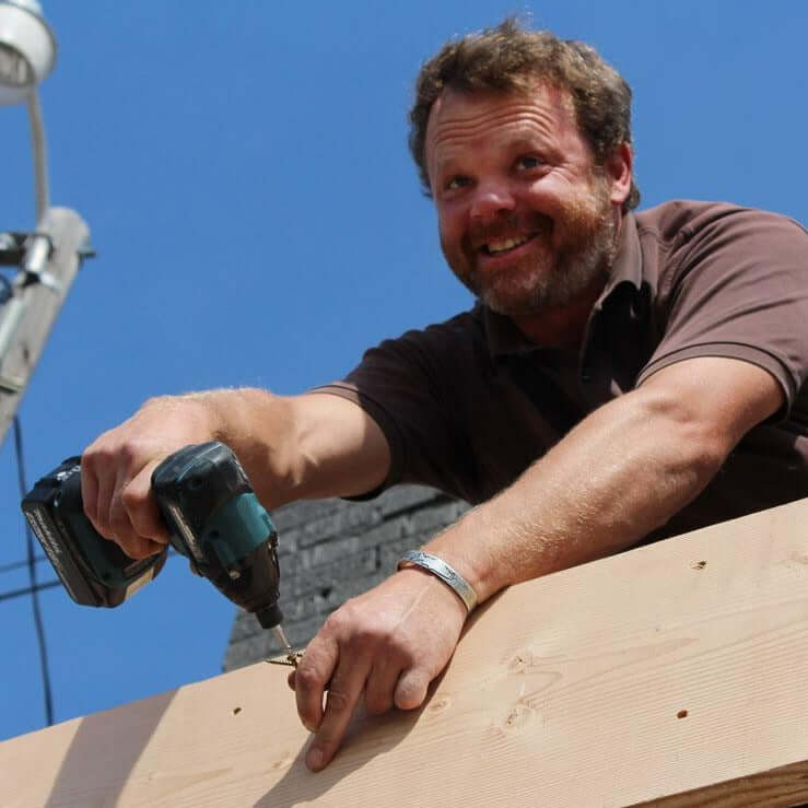 Man using a drill to secure a wooden board outdoors, showcasing skilled craftsmanship and a happy demeanor.