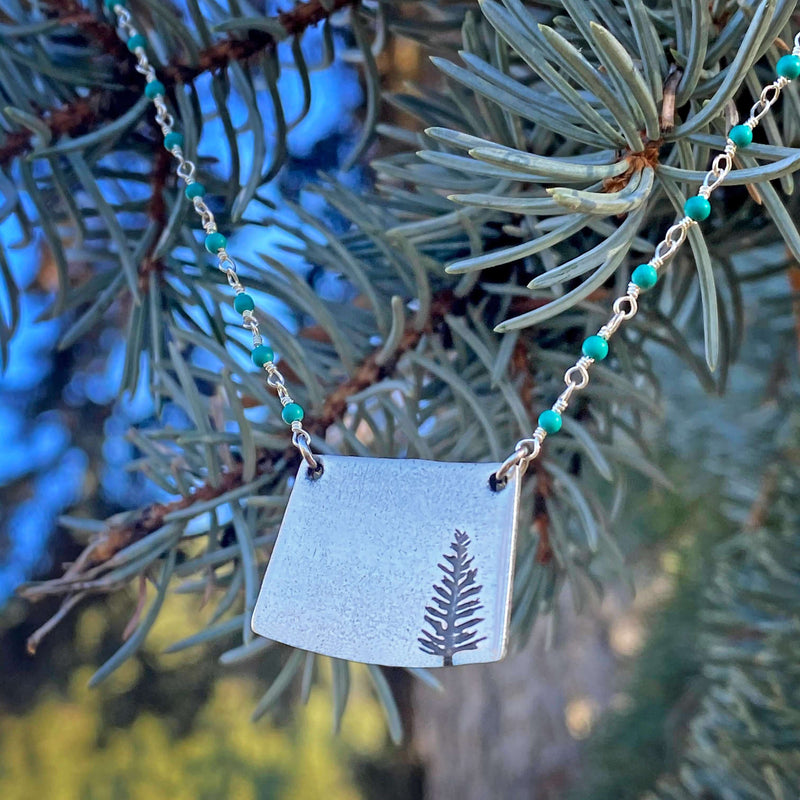 Spruce Necklace featuring a pendant with a tree design, set against a backdrop of evergreen branches.