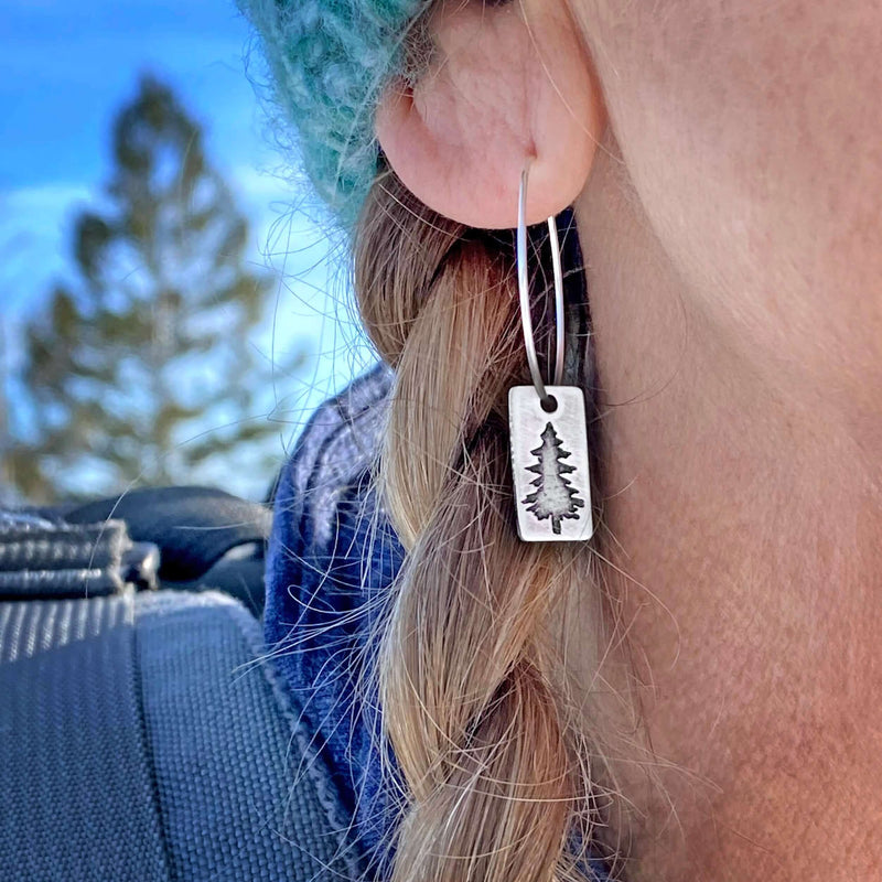 Spruce Tree Hoop Earrings worn by a person outdoors, featuring a small spruce tree design on a silver hoop earring.