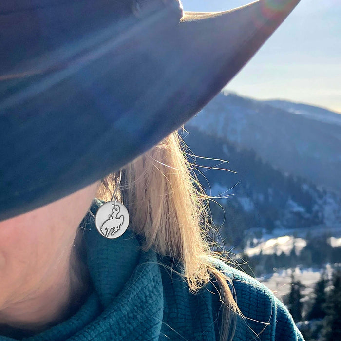 Close-up of a woman's ear wearing Steamboat Earrings against a mountain backdrop, showcasing the bronco design.