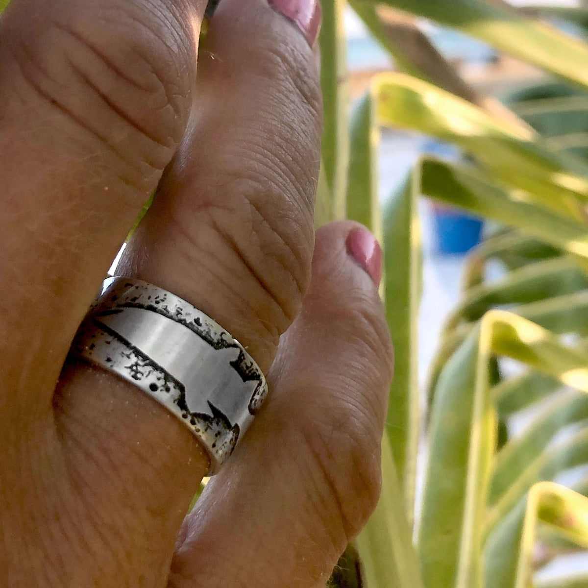 Hand showing a handmade Trout Ring with trout etching, surrounded by greenery, perfect for nature lovers.